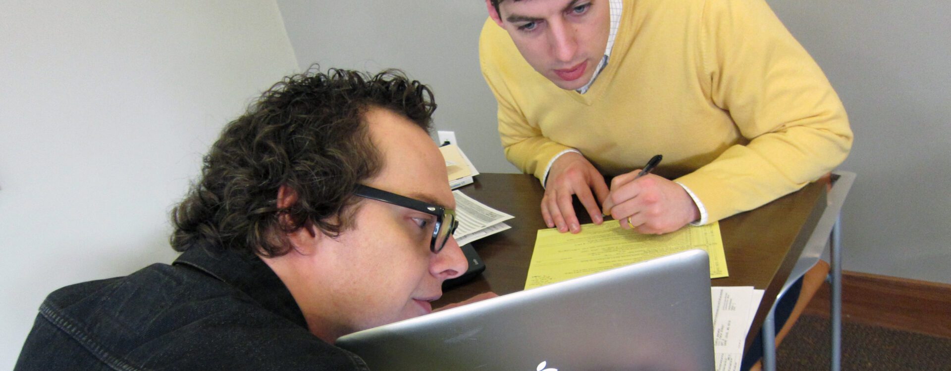 White artist with glasses wearing a black shirt holds a laptop while his white male volunteer, who is wearing a yellow sweater, makes notes and looks at the laptop screen