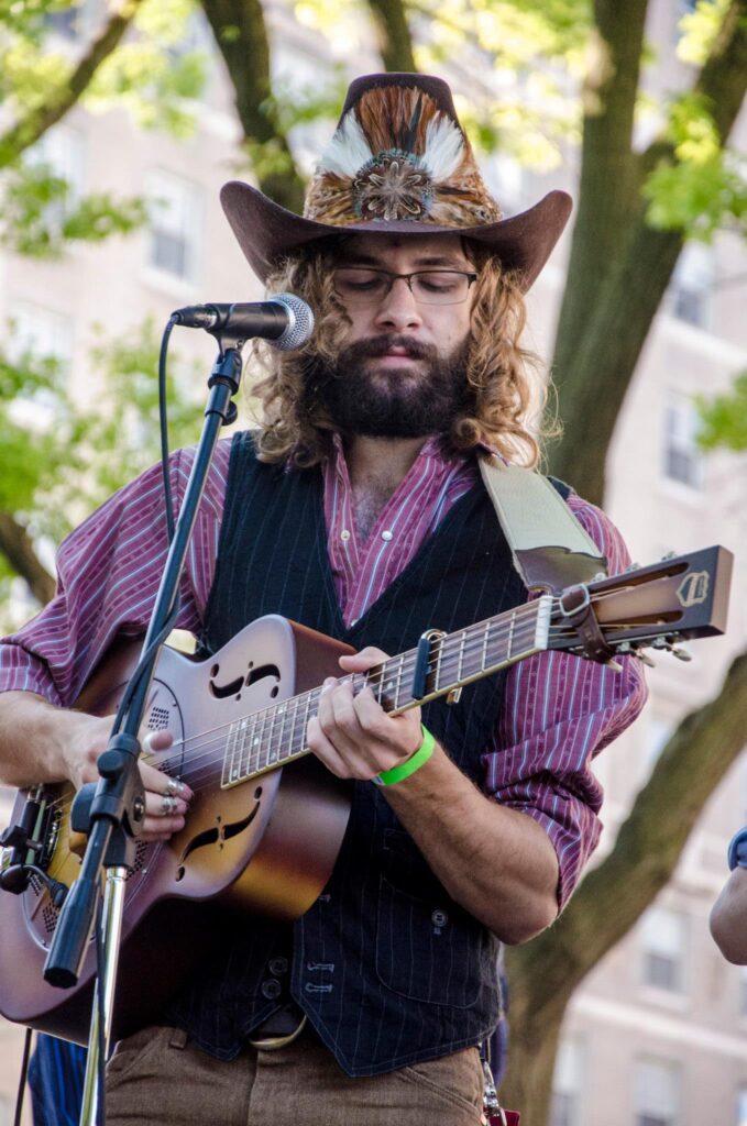 Nick Pence, white street performer wearing a cowboy hat, stripped purple shirt and black vest, playing his guitar