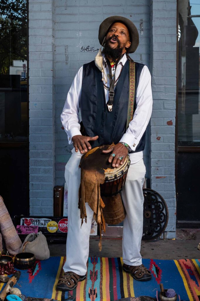 Raven Wolf C. Felton Jennings, a Black street performer, playing a drum and singing